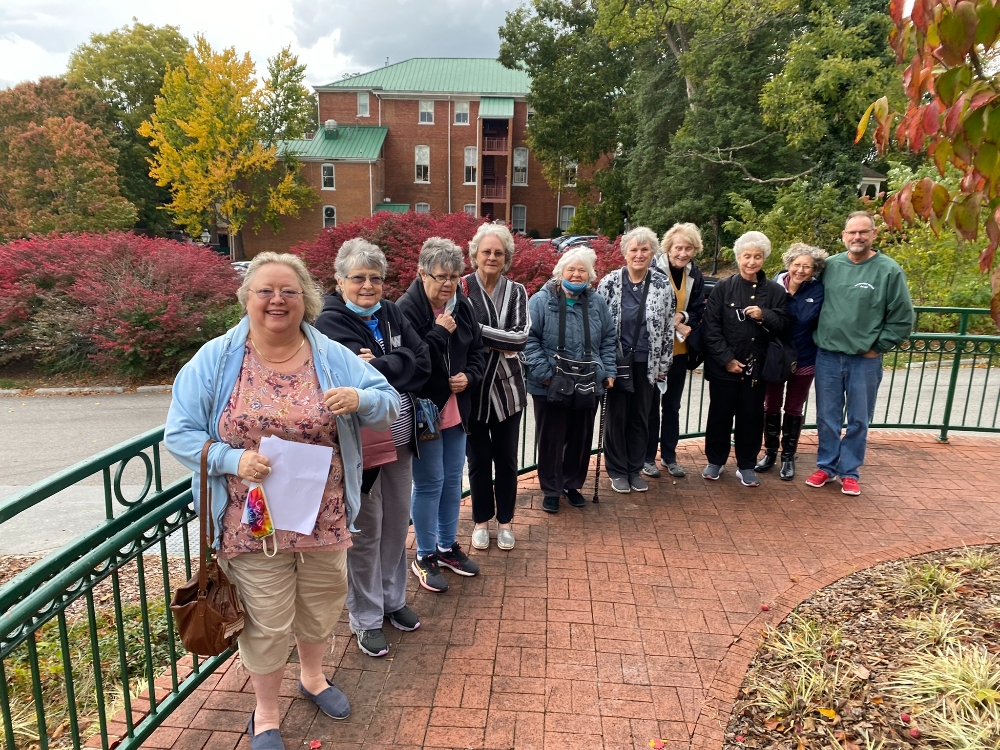 A group of adults pose for a picture along a curving brick sidewalk and green fence before heading to the Barter Theatre. The leaves on trees and shrubs are transitioning to fall with a mix of green, red, yellow, and orange leaves.